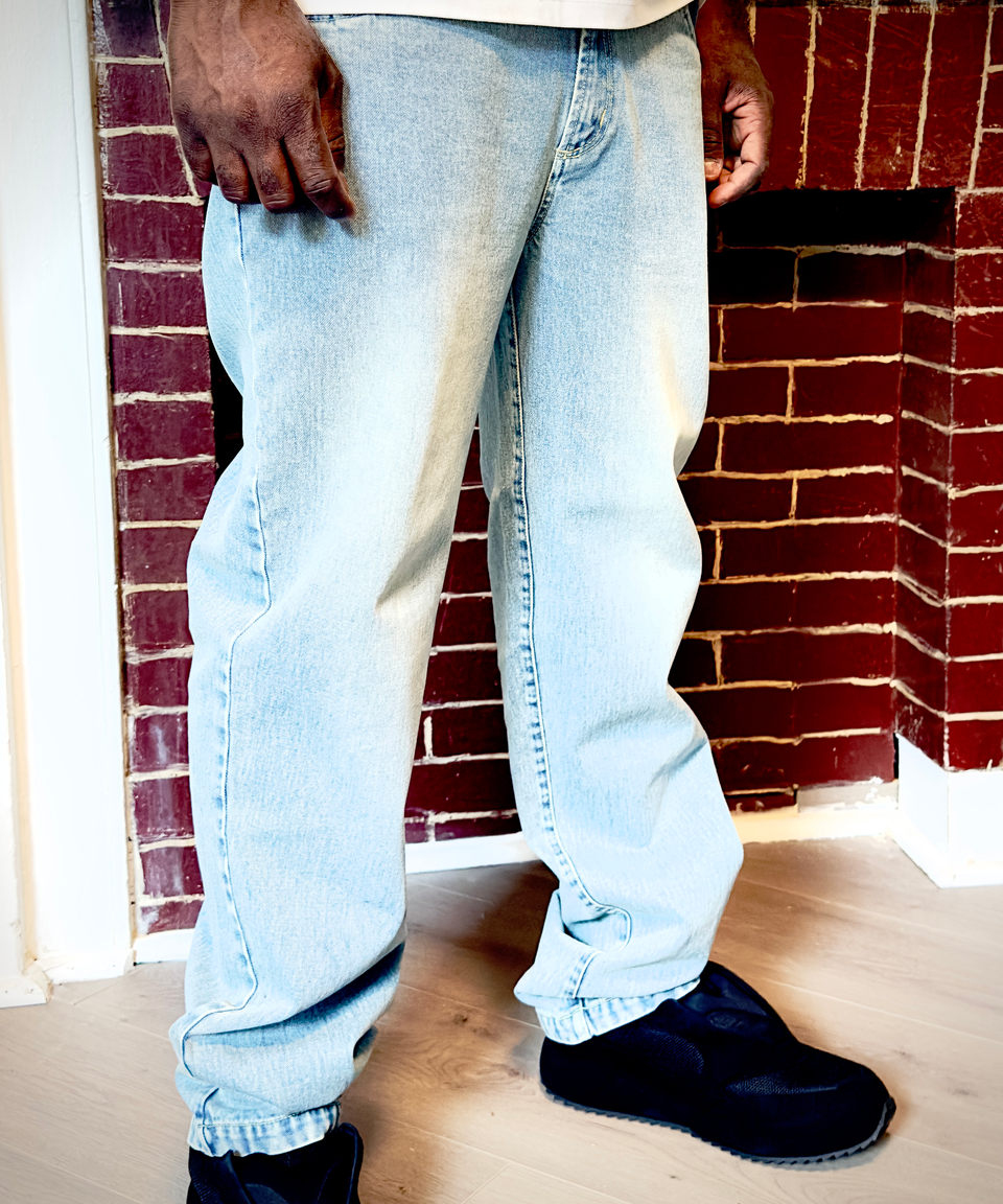 Person wearing light blue jeans and black shoes standing in front of a brick wall.