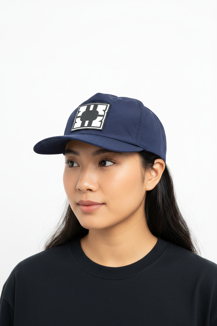 A woman wearing a navy blue nylon cap with a white and black PVC logo patch on the front, paired with a black crewneck shirt, against a plain white background.